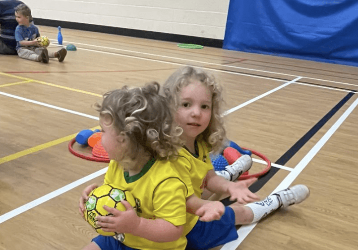 Best classes for toddlers. Image shows two toddlers sitting back to back, one holding a Socatots football.