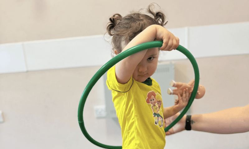Activities for toddlers near me. Image shows a young girl with a hulahoop round her and holding a baby doll.